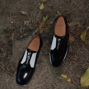 Puede incluir: Un par de zapatos de cuero negro con detalles en cuero blanco y una cremallera en la parte delantera. Los zapatos tienen una punta redonda y un tac&oacute;n bajo. Los zapatos est&aacute;n sobre un fondo marr&oacute;n con hojas.