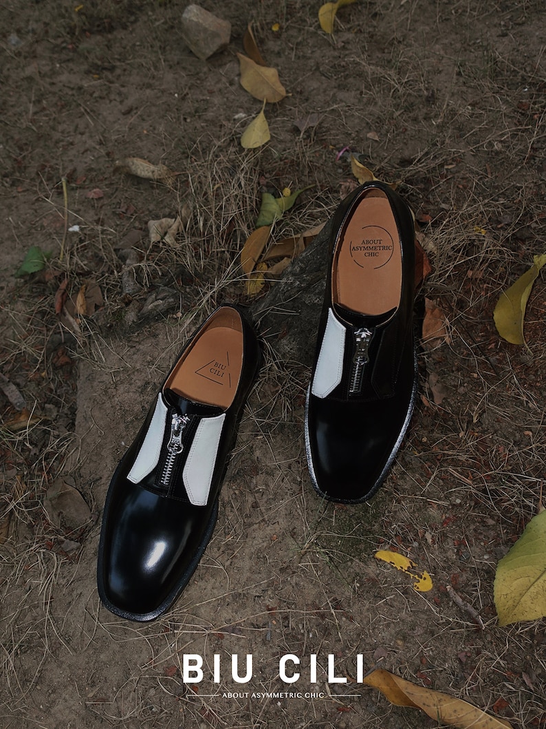 Puede incluir: Un par de zapatos de cuero negro con detalles en cuero blanco y una cremallera en la parte delantera. Los zapatos tienen una punta redonda y un tac&oacute;n bajo. Los zapatos est&aacute;n sobre un fondo marr&oacute;n con hojas.