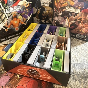 May include: A black box with a red and orange design. The box has multiple compartments for storing cards and game pieces. The box is labeled "Battle of Legends" and "Unmatched".