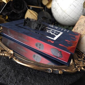 May include: A dark blue book with the word "MOUTH" and a silver design on the cover. The edges are red with skull and blood drop designs. A black sculptural element sits atop the book, which is reflected in a gold-rimmed mirror.