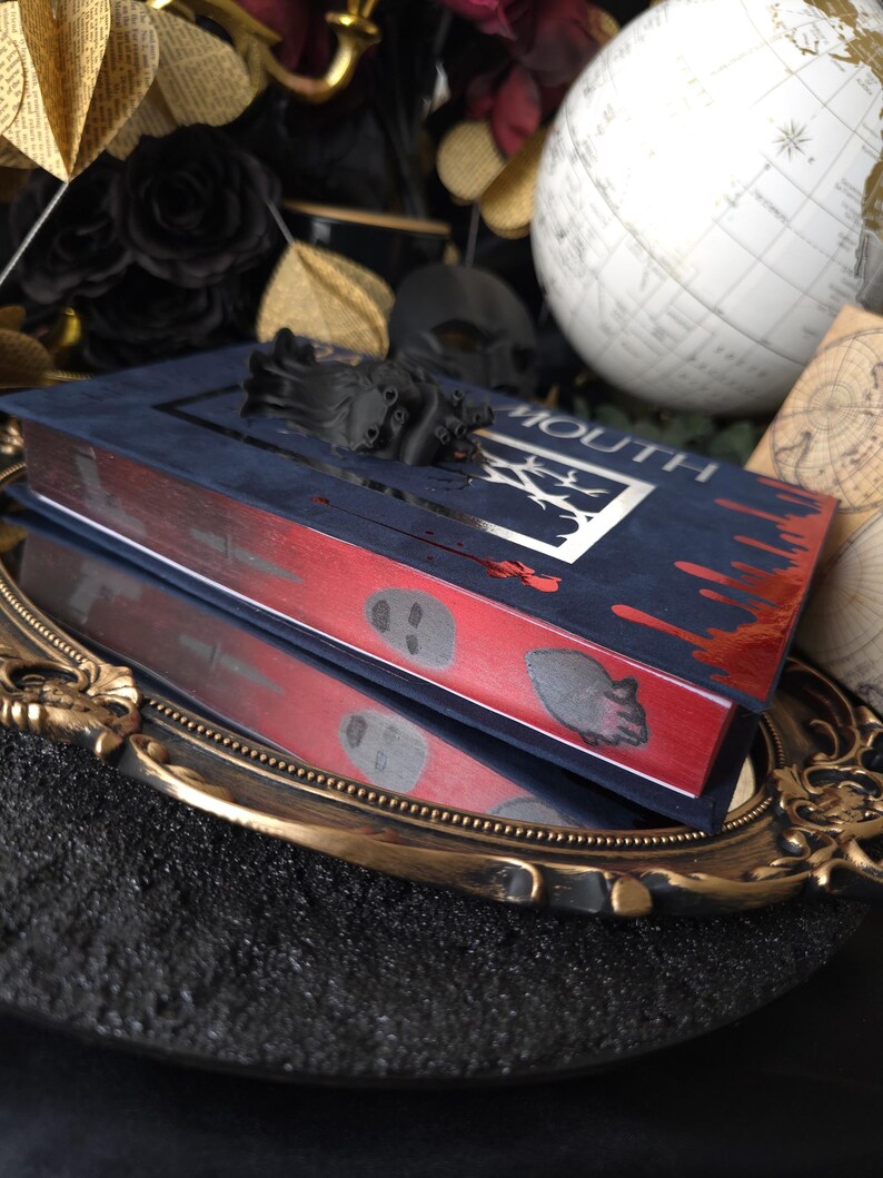 May include: A dark blue book with the word "MOUTH" and a silver design on the cover. The edges are red with skull and blood drop designs. A black sculptural element sits atop the book, which is reflected in a gold-rimmed mirror.