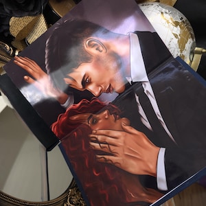 May include: Two books with illustrations of a man in a black suit and a woman with red hair. The man is leaning over the woman, with his hand covering her mouth. The books are on a dark surface with decorative elements.