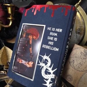 May include: A dark blue book with a silver frame and a picture of two people. The book has the text "HE IS HER RUIN. SHE IS HIS REBELLION" and a silver design. The top edge has a red, dripping design.