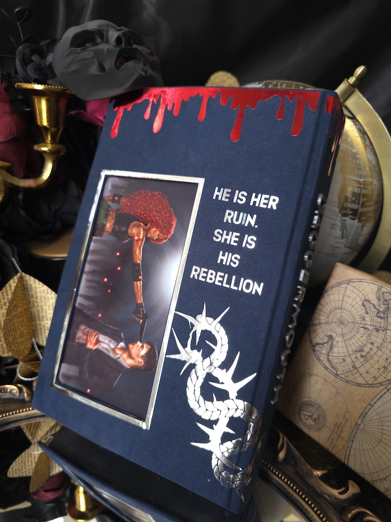 May include: A dark blue book with a silver frame and a picture of two people. The book has the text "HE IS HER RUIN. SHE IS HIS REBELLION" and a silver design. The top edge has a red, dripping design.