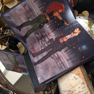 May include: Open book with a dramatic illustration of a woman with red hair pointing a gun at a man in a suit. The scene is set in a dark, urban environment with red accents. The book is open, revealing the artwork.