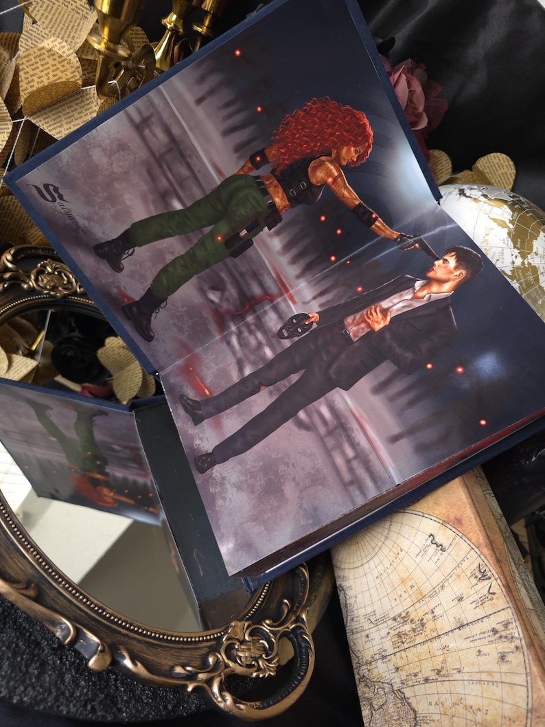 May include: Open book with a dramatic illustration of a woman with red hair pointing a gun at a man in a suit. The scene is set in a dark, urban environment with red accents. The book is open, revealing the artwork.