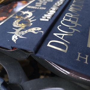 May include: Close-up of a navy blue book with silver lettering and a decorative snake design. The book's title, "Dagger Mouth," is prominently displayed. Additional text includes "He is her run, she is his rebellion."