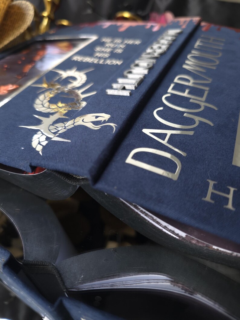 May include: Close-up of a navy blue book with silver lettering and a decorative snake design. The book's title, "Dagger Mouth," is prominently displayed. Additional text includes "He is her run, she is his rebellion."