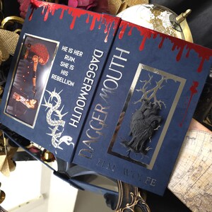 May include: A hardback book titled "Dagger Mouth" with a navy blue cover and gold lettering. The top edge has a red blood-like design. The book features a decorative illustration and the text "He is her ruin. She is his rebellion."