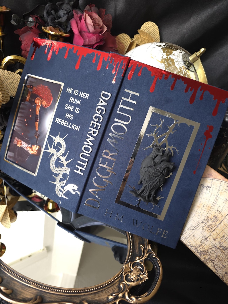 May include: A hardback book titled "Dagger Mouth" with a navy blue cover and gold lettering. The top edge has a red blood-like design. The book features a decorative illustration and the text "He is her ruin. She is his rebellion."