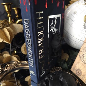 May include: A dark blue book titled "Dagger Mouth" with gold lettering and a red, dripping blood design on the top edge. The book is propped up by a black decorative stand, with a globe and other decorative items in the background.