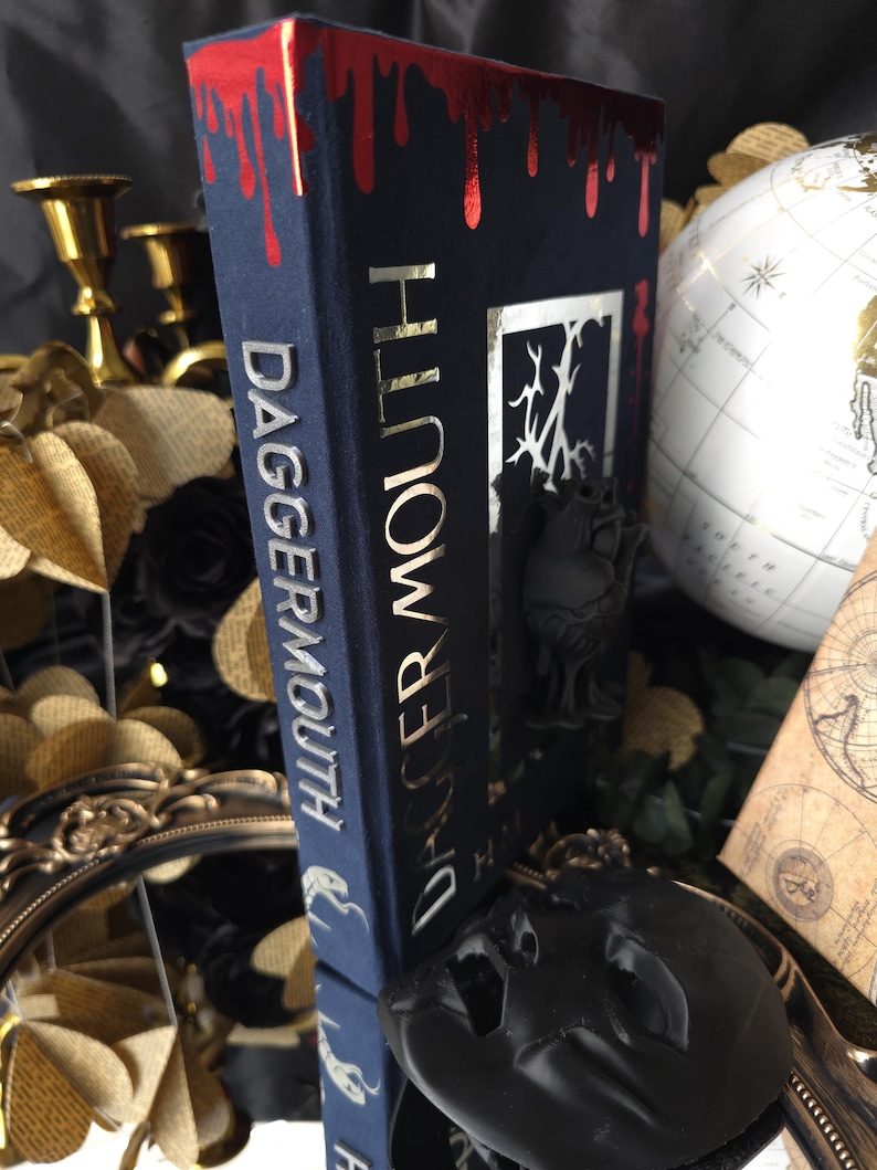May include: A dark blue book titled "Dagger Mouth" with gold lettering and a red, dripping blood design on the top edge. The book is propped up by a black decorative stand, with a globe and other decorative items in the background.