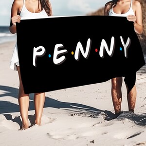 May include: Two people on a beach holding a black beach towel with the text "P.E.N.N.Y." in white letters with colorful dots.