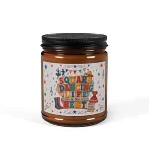 May include: A brown glass candle jar with a black lid featuring a label with the text SQUARE DANCING LIFE is the BEST and LIVE LAUGH DANCE. The label includes illustrations of cowboy boots, a hat, a dress, bunting, and musical notes.