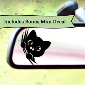 May include: A black cat decal peeking from behind a car mirror. The cat has large, round eyes and is holding onto the mirror with its paws. The text "Includes Bonus Mini Decal" is above the cat.