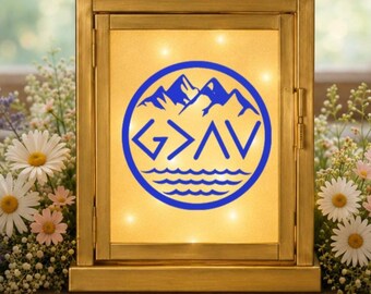 May include: A rectangular gold-toned lantern with a frosted glass panel featuring a blue circular graphic. The design includes mountain peaks above the text G>AV and wavy lines representing water at the bottom. The lantern is illuminated from within.