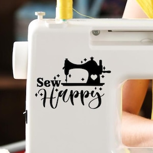 May include: A white sewing machine with a black decal that reads "Sew Happy." The decal features a silhouette of a sewing machine with a heart and star accents. Yellow thread is visible on top of the machine.