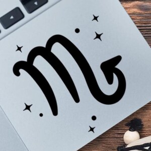 Puede incluir: Una calcomanía de vinilo negro con el símbolo del zodiaco Escorpio, caracterizado por la letra M con una cola curva y puntiaguda. El diseño está acentuado con pequeñas estrellas de cuatro puntas y puntos circulares alrededor del icono central.