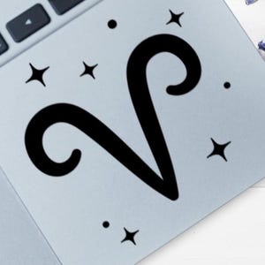 Puede incluir: Símbolo del zodiaco Aries negro con estrellas y puntos negros. El diseño está sobre una superficie azul claro, posiblemente una pegatina o calcomanía. La imagen está en un portátil.