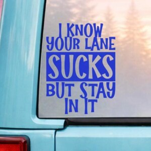 May include: A blue vinyl decal on a light blue vehicle window. The decal reads "I KNOW YOUR LANE SUCKS BUT STAY IN IT" in a bold, stylized font. The background shows a blurred forest scene.
