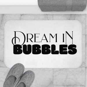 Könnte beinhalten: Weiße Badematte mit schwarzem Text, der "Dream in Bubbles" lautet.