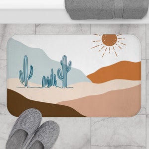 May include: A white bath mat with a desert landscape design. The design features a blue sky, brown mountains, a sun with rays, and blue cactus plants.