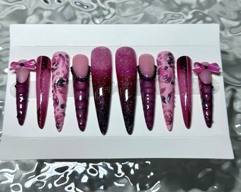 Peut inclure: Un ensemble de dix faux ongles longs en forme de stiletto. Les ongles présentent une variété de motifs, notamment des effets ombrés, des paillettes, des motifs floraux et des nœuds. Les couleurs incluent des nuances de rose, de violet et de noir, avec des perles noires.