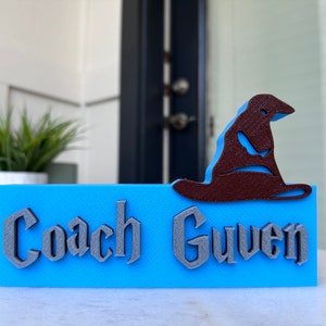 May include: A blue sign with the text "Coach Guven" in a gray, stylized font. A brown, 3D printed Sorting Hat sits on top of the sign.