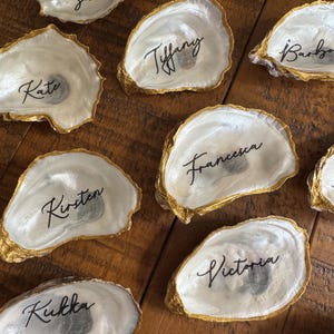 May include: A collection of oyster shells with gold-painted edges, each inscribed with a different name in elegant black script. The shells have a pearlescent white interior and are arranged on a wooden surface.