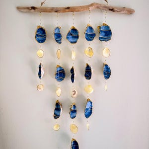 May include: A handcrafted wall hanging featuring a natural driftwood branch. Suspended from the branch are blue and gold-trimmed oyster shells and smaller, cream-colored shells, creating a coastal-inspired mobile. The shells are strung together with delicate wire.