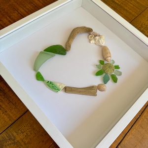 May include: A white frame with a heart-shaped arrangement of sea glass, driftwood, and seashells. The sea glass is green and the driftwood is brown. The seashells are white and beige.
