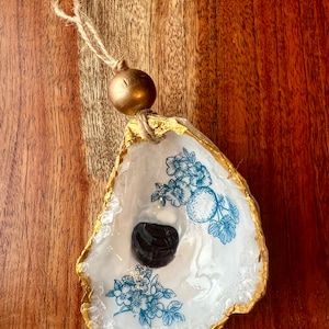 May include: A white oyster shell ornament with a blue floral design and a dark grey stone inside. The shell is accented with gold paint and has a gold bead on a twine hanger.