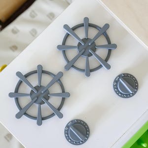 May include: A close-up of a white toy kitchen stove top with two gray burner grates and two gray control knobs. The knobs are labeled with numbers and the word "OFF".