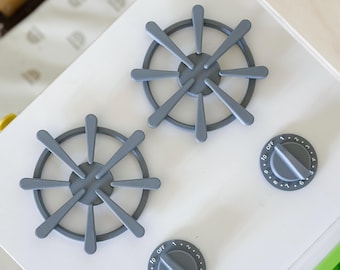 May include: A close-up of a white toy kitchen stove top with two gray burner grates and two gray control knobs. The knobs are labeled with numbers and the word "OFF".