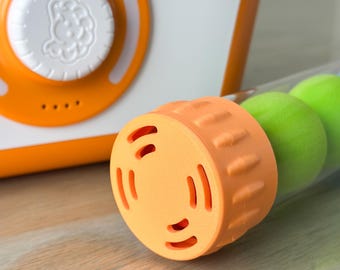 May include: A white and orange plastic device with a circular dial featuring a small embossed character icon, positioned next to a clear cylindrical tube containing two bright green spheres and capped with a textured orange plastic end piece.