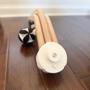 May include: A wooden baby activity gym frame featuring a light-colored natural wood arch structure with white circular end caps. A black and white patterned fabric ball hangs from the frame, providing a high-contrast visual element.
