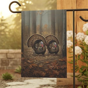 May include: A garden flag featuring a detailed painting of two turkeys in a forest setting. The turkeys have brown and black feathers, red wattles, and are surrounded by fallen leaves. The flag is displayed on a black metal stand.