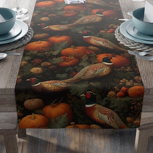Puede incluir: Un camino de mesa oscuro con un estampado detallado de calabazas, faisanes y follaje, ideal para una mesa de comedor. El camino de mesa se muestra sobre una mesa de madera con cubiertos y un jarrón con flores rosas. El diseño evoca el otoño.
