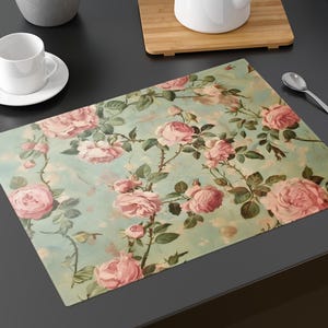 May include: A rectangular placemat with a vintage floral design featuring pink roses and green leaves on a light blue background. A white teacup and saucer, a teapot, and a spoon are also visible.