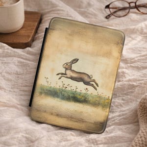 Puede incluir: Una funda para tableta de color beige con una ilustración vintage de una liebre corriendo. La obra de arte representa a la liebre en pleno salto sobre un campo de flores silvestres. La funda tiene un aspecto envejecido, lo que sugiere un estilo clásico o antiguo.