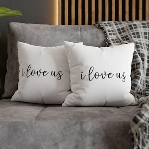 Könnte beinhalten: Zwei weiße Kissen mit dem Text "i love us" in schwarzer Schrift auf einem grauen Sofa.