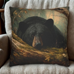Op de afbeelding: Decoratief sierkussen met een gedetailleerd schilderij van een zwarte beer die rust op een mosachtige boomstam. De beer heeft donkere vacht en een bruine snuit. De achtergrond toont een boslandschap met gedempte groen- en bruintinten.