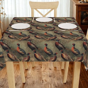 Puede incluir: Un mantel rectangular con un patrón repetido de faisanes en un entorno naturalista. Las aves tienen plumaje rojo, marrón y azul. Platos blancos están colocados sobre la mesa, que está rodeada de sillas de madera.
