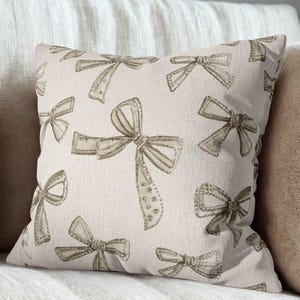 May include: A square decorative pillow with a repeating pattern of beige bows on a cream-colored background. The bows have a textured appearance and are embellished with small, sparkling details. The pillow is placed on a beige sofa.