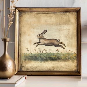 May include: A framed painting of a brown rabbit hopping through a field of green grass and yellow flowers. The frame is a rustic gold color.