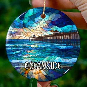 Könnte beinhalten: Runder Keramikschmuck mit einer Darstellung eines Piers über dem Meer im Glasmalerei-Stil. Der Schmuck hat eine goldene Aufhängeschnur und den weißen Schriftzug "OCEANSIDE".
