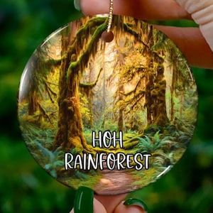May include: A round Christmas ornament featuring a vibrant rainforest scene with the text "HOH RAINFOREST". The ornament has a gold string for hanging and is held by a hand with green nail polish.