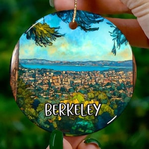 May include: A circular ceramic Christmas ornament depicting a landscape of Berkeley, California, with a blue sky and water. The word "BERKELEY" is printed in white. It has a gold hanging string.