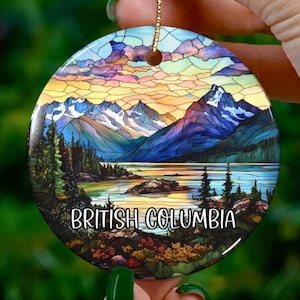 Puede incluir: Un adorno redondo de cerámica con un diseño de vitral de una escena de lago de montaña. El adorno presenta un lago azul, árboles verdes y montañas cubiertas de nieve. El texto "British Columbia" está impreso en el adorno.