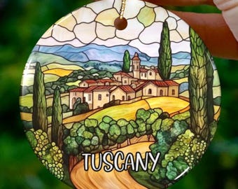 May include: A round ceramic ornament with a stained glass design of a Tuscan landscape. The ornament features a village with buildings, trees, and a winding road. The text "Tuscany" is written on the road.